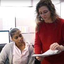 a woman in a red shirt goes over paperwork with an intern sitting at her computer